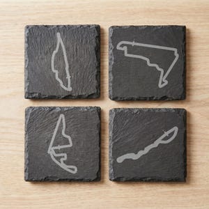 May include: A set of four square black slate coasters, each featuring a distinct light gray engraved outline of a different professional motor racing circuit track layout. The coasters have a rough, natural textured edge and a flat surface.
