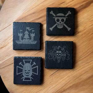 May include: Four square black slate coasters with engraved designs. One features a skull and crossbones, another a castle, and two others with unique skull and crossbones designs. The coasters are arranged on a wooden surface.