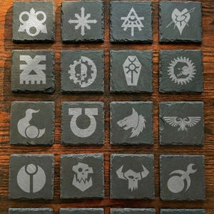 May include: A collection of twenty square, dark gray slate coasters, each etched with a different symbol. The symbols are in a lighter gray and include various emblems, logos, and stylized designs. The coasters are arranged on a wooden surface.