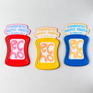May include: Three colorful magnetic photo frames for Instax Mini photos. The frames are red, yellow, and blue, each with a matching top banner that reads "MAGNETIC PHOTO FRAME". The frames have a unique wavy design and the word "echo" in the center.