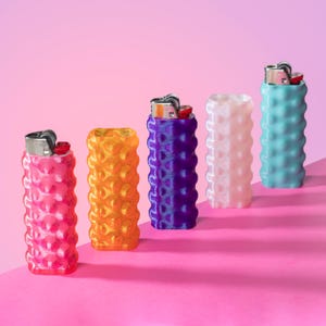 May include: Five colorful lighter cases in pink, orange, purple, white, and teal. Each case has a textured, rounded pattern. The lighters inside have silver metal tops and red ignition buttons, set against a pink background.
