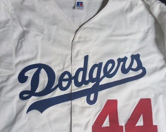 May include: White baseball jersey with the word "Dodgers" in blue script across the chest, and the number "44" in red. The jersey has a V-neck and button-up front. A small tag is visible near the collar.