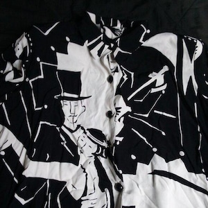 May include: Black and white button-down shirt with a unique graphic design. The shirt features a repeating pattern of figures in hats and abstract shapes. The collar and button placket are white, while the rest of the shirt is black.