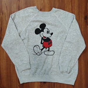 May include: A gray sweatshirt featuring a classic Mickey Mouse graphic. The cartoon character is depicted in black, red, and white. The sweatshirt has long sleeves and a crew neck.