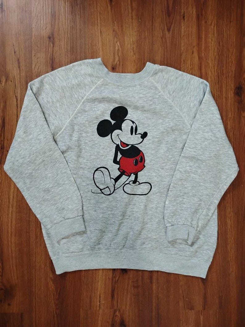 May include: A gray sweatshirt featuring a classic Mickey Mouse graphic. The cartoon character is depicted in black, red, and white. The sweatshirt has long sleeves and a crew neck.