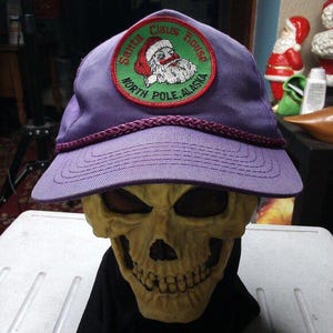 Può includere: Un cappellino da baseball viola con visiera curva e bordo decorativo in corda. Il cappellino presenta una toppa circolare con le parole "Santa Claus House, North Pole, Alaska" e un'immagine di Babbo Natale.