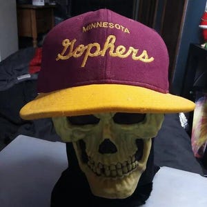 Może przedstawiać: Bordowa i złota czapka bejsbolowa z napisem "Minnesota Gophers" wyszytym złotą nicią. Czapka ma żółty daszek i jest prezentowana na czaszce.