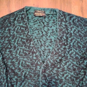 Op de afbeelding: Een middelgroot, teal en zwart patroon vest met een V-hals en een enkele knoopsluiting. Het label luidt "UNIFORM CODE". De trui heeft een textuur gebreid ontwerp.