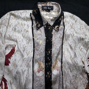 Può includere: Una camicia bianca a maniche lunghe con bottoni e un motivo a righe nere e dorate. La camicia ha un colletto e una chiusura con bottoni sul davanti. La camicia ha un'etichetta che dice "Silk Boy".