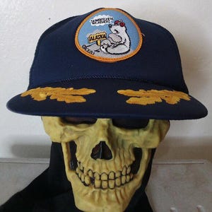 May include: A navy blue trucker hat with a yellow embroidered patch featuring a polar bear and the text "Snarflichen Weather! Alaska".