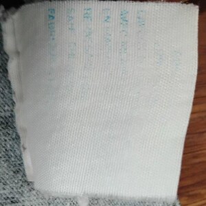 May include: A white fabric label with light blue text. The label is rectangular and attached to a grey fabric. The text is partially visible, with words like "FABRIC" and other details.