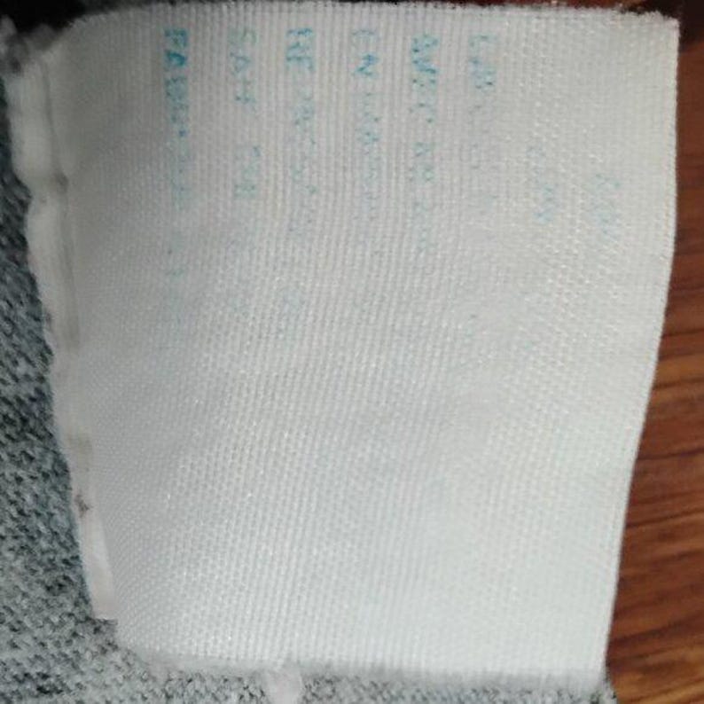 May include: A white fabric label with light blue text. The label is rectangular and attached to a grey fabric. The text is partially visible, with words like "FABRIC" and other details.