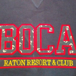 May include: Navy blue sweatshirt with the word "BOCA" in large red letters outlined in red. The letters are filled with a pattern of white anchors. Below the word "BOCA" is the text "RATON RESORT & CLUB" in yellow.