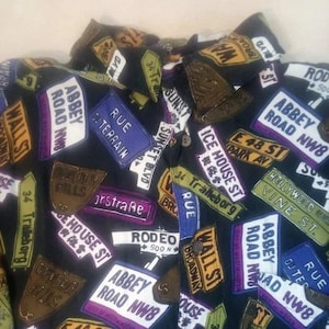 May include: Black button-up shirt with a street sign pattern. The signs feature various street names, including Abbey Road NW8, Wall St, Rodeo, and Ice House St.