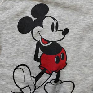 May include: A gray sweatshirt featuring a large graphic of Mickey Mouse. The cartoon character is depicted in black, white, and red, with a smiling expression and his signature red shorts. The sweatshirt has a crew neck.
