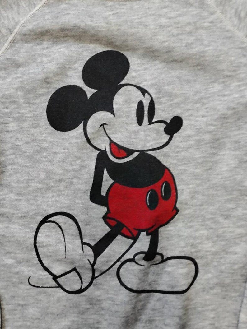 May include: A gray sweatshirt featuring a large graphic of Mickey Mouse. The cartoon character is depicted in black, white, and red, with a smiling expression and his signature red shorts. The sweatshirt has a crew neck.