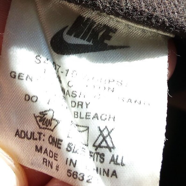 K&ouml;nnte beinhalten: Wei&szlig;es Pflegeetikett mit schwarzem Text und Symbolen. Das Etikett enth&auml;lt das Nike-Logo, Waschanweisungen und den Text "Adult: One Size Fits All", "Made in China" und "RN # 5632".