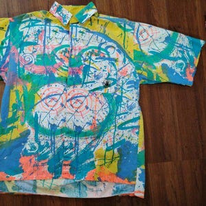 Puede incluir: Camisa de manga corta con botones y un llamativo dise&ntilde;o abstracto. La camisa presenta un colorido patr&oacute;n de salpicaduras y pinceladas turquesas, amarillas, naranjas y blancas. Tiene cuello y un peque&ntilde;o logotipo en el pecho.