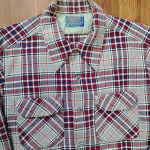 Può includere: Una camicia a maniche lunghe abbottonata con un motivo a quadri nei toni del grigio, rosso e blu navy. La camicia presenta un colletto a punta, due tasche frontali sul petto con patta e un'etichetta del marchio Pendleton sul colletto.