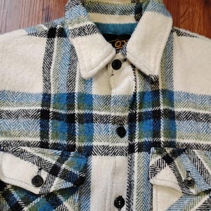 Peut inclure: Une chemise à carreaux blancs, bleus et noirs avec un col et deux poches de poitrine boutonnées. La chemise a des boutons noirs et un tissu texturé. Le motif à carreaux est un mélange de bleu, noir et vert sur fond blanc.