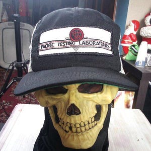May include: A black trucker hat with a white patch featuring the text "PACIFIC TESTING LABORATORIES" in black and red. The hat is worn over a yellow skull mask with dark eye sockets and a wide grin. The hat has a black brim and a white mesh back.