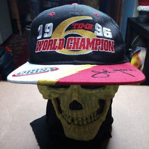 May include: Black and red baseball cap with a white brim. The cap has a gold embroidered logo that says "1996 Time World Champion". The cap is signed in black ink.