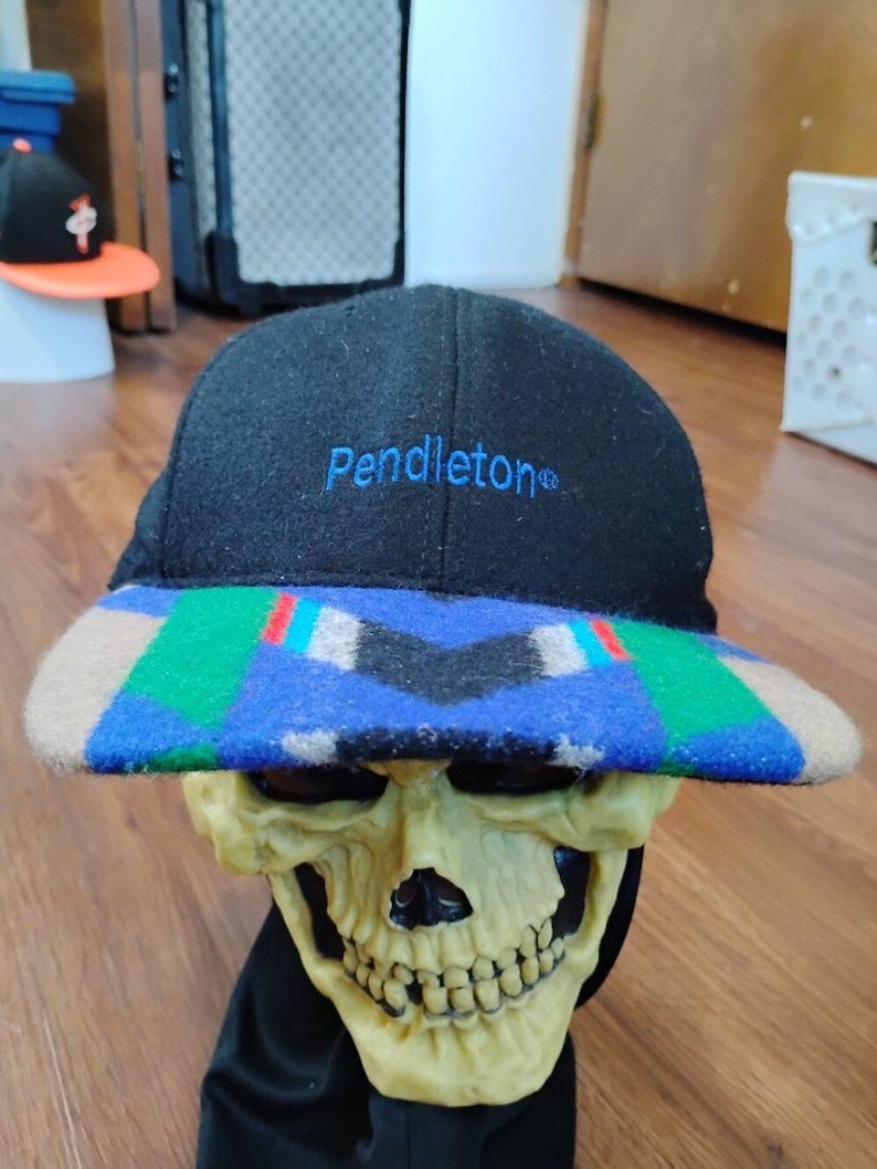 May include: A black baseball cap with the word Pendleton embroidered in blue thread on the front. The brim features a geometric pattern in shades of blue, green, tan, black, and red. The cap is made of a felted material and has a structured crown.