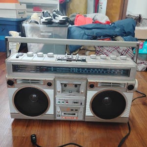 Puede incluir: Un radiocassette portátil Audiovox de color plateado con dos altavoces negros, una platina de casete central y un sintonizador de radio AM/FM con pantalla de frecuencia horizontal. El dispositivo incluye un asa de transporte, varios mandos de control plateados y un cable de alimentación negro.