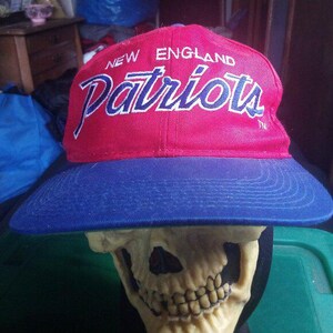 May include: A red and blue New England Patriots baseball cap. The cap features the team name "Patriots" in white script across the front, with "New England" above it. The brim is blue, and the cap is set against a blurred background.