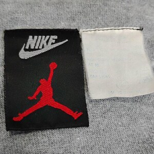 May include: A close-up of a black rectangular tag featuring the white Nike logo and a red Jumpman logo. A white tag is attached to the right. The background is grey fabric.