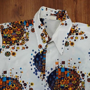 May include: White button-up shirt with a bold, colorful pattern. The design features a mix of orange, yellow, blue, and brown circles and squares. The shirt has a classic collar and button closure, perfect for a retro or vintage look.