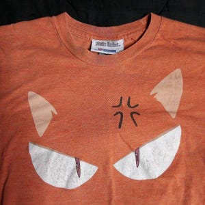 May include: Orange t-shirt featuring a cartoon face design. The design includes white eyes with purple accents, white ears, and a black symbol above the eyes. The shirt has a crew neck and a tag at the neckline.