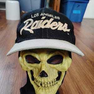 以下が含まれることがあります： ベージュの「LOS ANGELES Raiders」が刺繍された黒い野球帽。 つばはベージュで、黄色のスカルマスクに飾られています。 ヴィンテージスタイル。