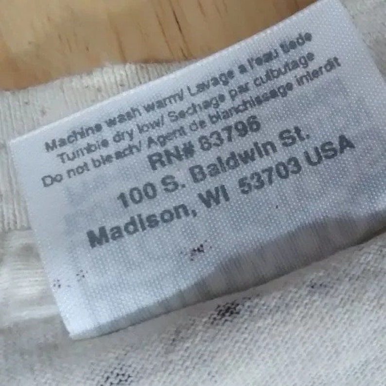 May include: White clothing care label with instructions for washing and drying. The label also includes the company's registration number, address, and location: RN# 83796, 100 S. Baldwin St., Madison, WI 53703 USA.