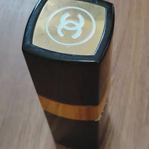 May include: A rectangular lipstick tube with a glossy black finish and a gold-colored band around the center. The top cap features a square gold-colored inlay with a white interlocking double C logo centered on the surface of the lid.