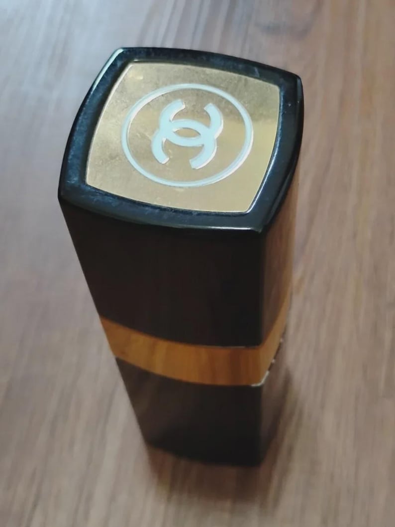 May include: A rectangular lipstick tube with a glossy black finish and a gold-colored band around the center. The top cap features a square gold-colored inlay with a white interlocking double C logo centered on the surface of the lid.