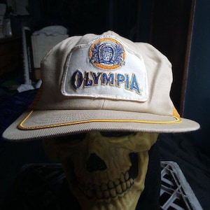 May include: A beige baseball cap with a white patch displaying the word "OLYMPIA" in blue. The patch also features a circular emblem with blue and gold details. The cap has a yellow trim.