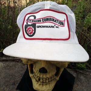 Puede incluir: Gorra de camionero blanca con parte trasera de malla y ala ancha. El parche frontal presenta el texto "THE WINNING COMBINATION" y "GROWMARK INC." en rojo y negro. El sombrero se muestra sobre una calavera.