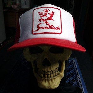 Puede incluir: Gorra de camionero roja y blanca con la palabra "Snowbirds" bordada en la parte delantera, colocada sobre una calavera amarilla. La gorra tiene una visera roja y un panel frontal blanco con un logotipo rojo.
