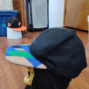 May include: A black baseball cap with a flat brim featuring a geometric pattern in blue, green, and tan. The cap is displayed on a skull-shaped stand. The crown is solid black with small ventilation eyelets and a blue embroidered detail on the side.