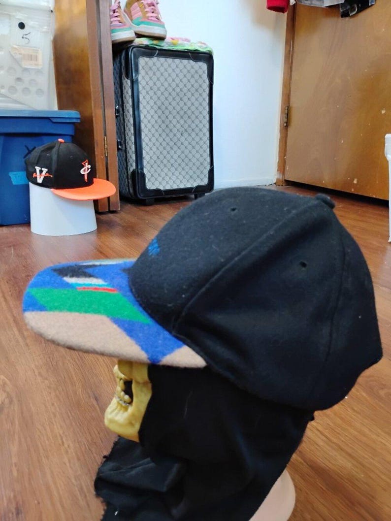 May include: A black baseball cap with a flat brim featuring a geometric pattern in blue, green, and tan. The cap is displayed on a skull-shaped stand. The crown is solid black with small ventilation eyelets and a blue embroidered detail on the side.