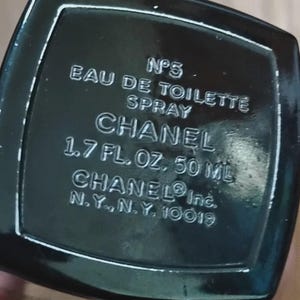May include: A black rectangular base of a perfume bottle with white embossed text reading N&deg;5 EAU DE TOILETTE SPRAY CHANEL 1.7 FL. OZ. 50 ML CHANEL Inc. N.Y., N.Y. 10019. The surface is glossy and dark, featuring clear, legible lettering throughout.