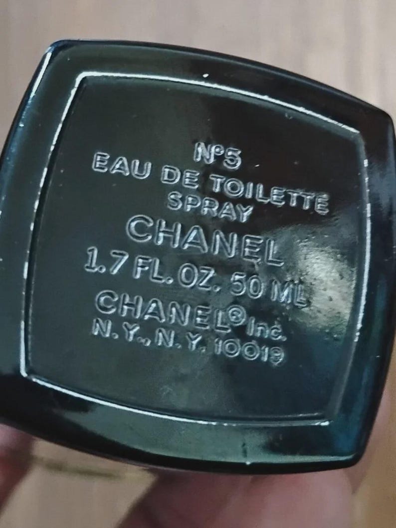 May include: A black rectangular base of a perfume bottle with white embossed text reading N&deg;5 EAU DE TOILETTE SPRAY CHANEL 1.7 FL. OZ. 50 ML CHANEL Inc. N.Y., N.Y. 10019. The surface is glossy and dark, featuring clear, legible lettering throughout.