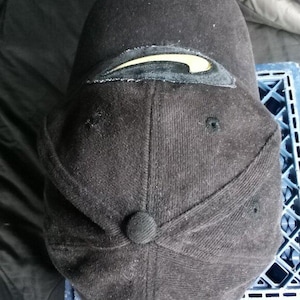 May include: Black baseball cap with a gold Nike swoosh logo. The cap has a curved brim and a button on the back for adjustment. The cap is made of a textured fabric.