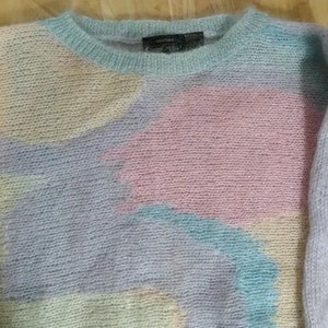 May include: A pastel-coloured fuzzy sweater with a light blue, pink, and yellow abstract pattern. The sweater has a crew neck and is made of a soft, fluffy material.