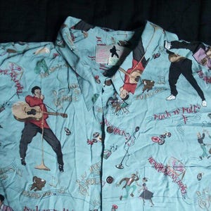 Puede incluir: Camisa celeste con botones con un patrón repetido de un músico tocando la guitarra, notas musicales y las palabras "Rock n' Roll". La camisa tiene cuello y mangas cortas. La camisa está hecha de una tela ligera.