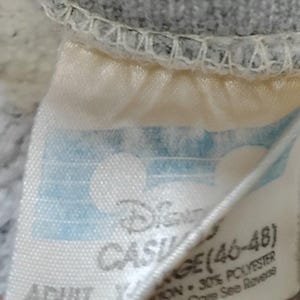 May include: Close-up of a tag from a gray sweatshirt. The tag is off-white with blue accents and features the Disney logo and the text "CASUAL AGE (46-48)". The tag also indicates the garment is adult size and lists the fabric content.