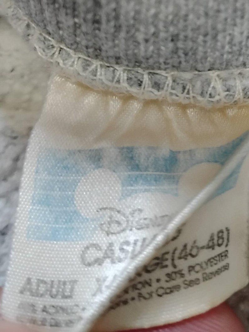 May include: Close-up of a tag from a gray sweatshirt. The tag is off-white with blue accents and features the Disney logo and the text "CASUAL AGE (46-48)". The tag also indicates the garment is adult size and lists the fabric content.