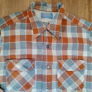 May include: A long-sleeve button-up shirt featuring a plaid pattern in shades of burnt orange, gray, and white. The shirt has a pointed collar, two front chest pockets with flaps, and a blue Pendleton brand label sewn inside the neck.