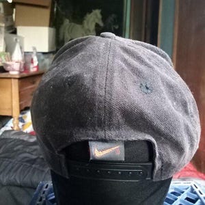 May include: A dark gray baseball cap with a black adjustable snapback closure. The Nike logo is visible on a small tag at the back. The cap has a rounded crown and a small button on top.